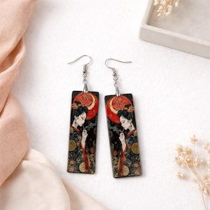 Könnte beinhalten: Ein Paar rechteckige Ohrhänger mit asiatisch inspiriertem Design. Jeder Ohrring zeigt eine Frau in einem traditionellen Kimono vor einem schwarzen Hintergrund mit einem roten Mond. Die Ohrringe haben silberfarbene Haken.
