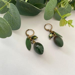 Puede incluir: Pendientes con temática de aceitunas. Cada pendiente presenta una aceituna verde oscuro, una pequeña hoja verde y cuentas verdes, suspendidas de un aro dorado. Los pendientes están sobre un fondo blanco con follaje verde.