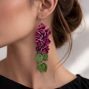 Op de afbeelding: Een lange oorhanger met een tros diep magenta gekleurde bloemen aan de bovenkant, die overgaat in een reeks gestructureerde groene bladeren aan de onderkant. De bloem- en bladelementen zijn bevestigd aan een zilverkleurige metalen haak.