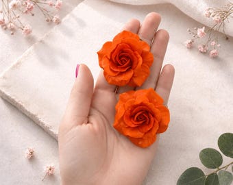 May include: Two vibrant orange rose-shaped embellishments with layered petals. The flowers feature a smooth, matte texture and are displayed resting in an open palm, showcasing their detailed, realistic floral structure and bright color.