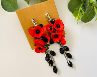 May include: Dangle earrings featuring clusters of vibrant red poppy flowers with black centers, accented by dangling black faceted beads on silver-toned metal chains. The earrings are attached to a square tan display card.