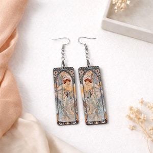 May include: A pair of rectangular dangle earrings featuring an Art Nouveau design. Each earring showcases a woman in a flowing gown amidst floral and botanical elements, with a color palette of beige, brown, and muted greens. The earrings have silver-tone hooks.