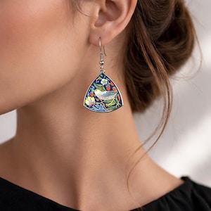 May include: Silver-tone dangle earrings with a triangular design. Each earring features a detailed illustration of a bird perched on a branch, surrounded by flowers and berries, set against a dark blue background. The earrings are a unique accessory.