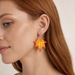 Op de afbeelding: Een goudkleurige oorring met een bungelende esdoornbladbedel. Het blad heeft een kleurverloop van levendig geel en oranje, met gedetailleerde nerven en gekartelde randen, hangend aan een dunne metalen ring die aan de oorlel is bevestigd.