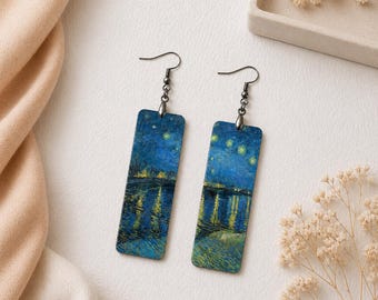 May include: A pair of rectangular drop earrings featuring a printed reproduction of a starry night landscape with deep blue tones, yellow light reflections, and dark metal hooks. The earrings have a flat, elongated shape with rounded corners.