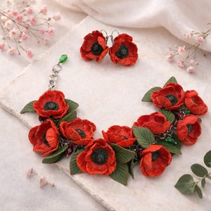 Può includere: Un set di gioielli composto da un bracciale e un paio di orecchini, ciascuno caratterizzato da fiori di papavero rossi scolpiti con centri scuri e foglie verdi. Il bracciale è realizzato con una catena in metallo argentato e perline verdi.