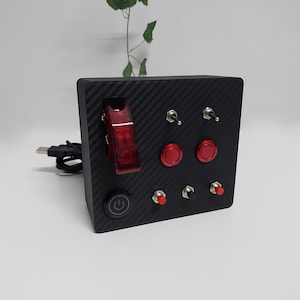 May include: A black box with a carbon fiber pattern and multiple buttons. There are two red toggle switches, two red push buttons, a black push button with a power symbol, and a red toggle switch with a clear plastic cover. The box is connected to a USB cable.