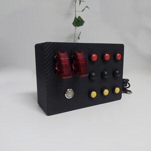 Pode incluir: Painel de controle preto com botões vermelhos e amarelos, duas chaves de alternância vermelhas e um botão prateado com a etiqueta "Iniciar motor".