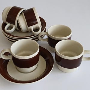 Op de afbeelding: Een set van vier crèmekleurige keramische kopjes en schoteltjes met een brede, horizontale donkerbruine band rond het midden van elk kopje en de rand van elk schoteltje. De kopjes hebben een afgerond oortje en een stapelbaar ontwerp.