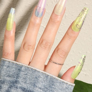 May include: A hand model wearing a denim jacket with long, light green and iridescent press-on nails with floral designs.