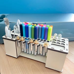 Könnte beinhalten: Ein cremefarbener Bastel-Organizer aus Holz mit mehreren Fächern. Er enthält verschiedene Bastelwerkzeuge, darunter Stifte, Marker in Regenbogenfarben und Schneidklingen. Der Hintergrund zeigt eine hellblaue Oberfläche.