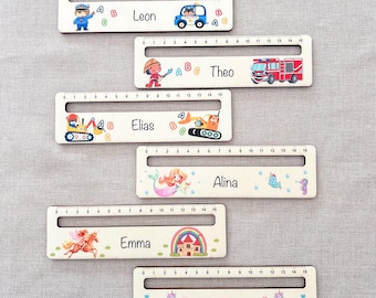 May include: Wooden rulers with names and illustrations. Each ruler features a name, a measurement scale up to 15 inches (approx. 38 cm), and a unique design, including a police car, fire truck, mermaid, and unicorn. The names are Leon, Theo, Elias, Alina, Emma, and Jessica.