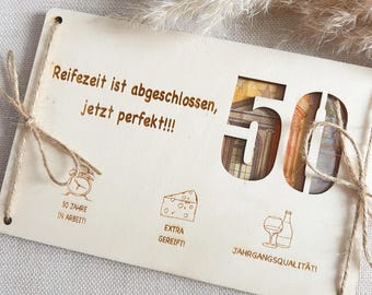 May include: Wooden card with the text "Reifezeit ist abgeschlossen, jetzt perfekt!!!" and the number 50. The card has a cutout for a 50 euro bill. There are also illustrations of a clock, a piece of cheese, and a glass of wine.