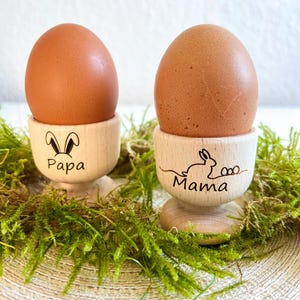 Könnte beinhalten: Zwei braune Eier in hölzernen Eierbechern. Ein Becher hat ein Hasenohren-Design und das Wort "Papa" darunter. Der andere Becher hat ein Hasen-Design und das Wort "Mama" darunter. Die Becher stehen auf grünem Moos.