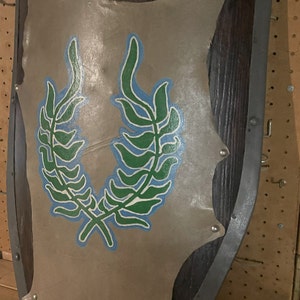 May include: A wooden shield with a metal rim and a gray leather surface. The shield has a green and blue laurel wreath design painted on the front.
