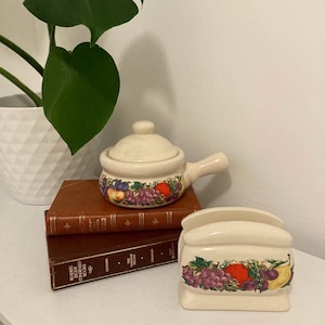 May include: A set of two ceramic kitchenware pieces featuring a fruit design. The pieces are a small pot with a lid and a napkin holder. Both pieces are white with a colourful design of grapes, apples, pears, and a red pepper.