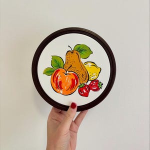May include: Round decorative plate with a white centre and a dark brown rim. The plate features a colourful illustration of a pear, apple, lemon, and strawberries with green leaves. A hand is holding the plate.