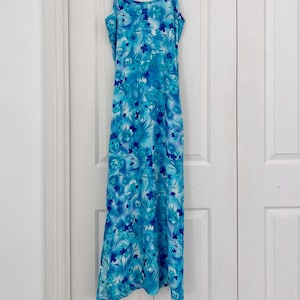 May include: A long, flowing dress with a blue floral pattern. The dress has thin spaghetti straps and a flared skirt. The fabric has a watercolour effect with shades of light and dark blue. The dress is hanging on a white hanger.