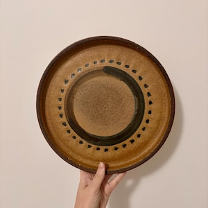 May include: A round ceramic plate in shades of brown and tan. It has a dark brown rim and a central design of a dark green semi-circle and black dots. The plate is held by a hand.