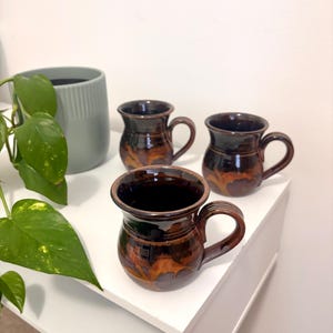 May include: Three ceramic mugs with a rounded, bulbous shape and a glossy finish. The mugs feature a dark brown and amber marbled glaze pattern, each with a curved handle and a flared rim. The interior of each mug is a solid, dark brown colour.