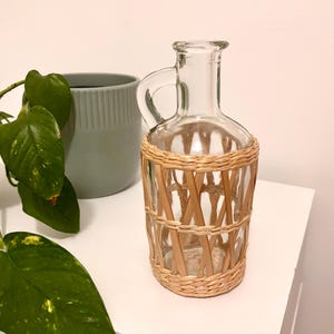 May include: A clear glass carafe with a narrow neck and a single handle, featuring a decorative woven light brown wicker wrap around the lower body. The glass is transparent, and the wicker has a crisscross pattern with horizontal bands.
