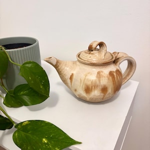 May include: A handcrafted ceramic teapot with a lid, finished with a beige and brown glaze. The teapot has a curved spout and handle. A green plant in a grey pot is visible in the background.
