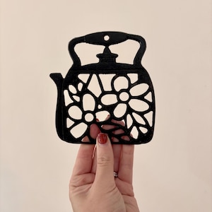 May include: Black metal teapot-shaped trivet with a floral cutout design. The trivet has a handle and a spout. The background is a solid, light beige colour. The trivet is held by a hand.