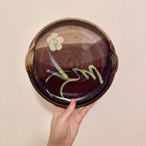 May include: A round ceramic plate with a dark brown and purple glaze. The plate features a white flower and abstract yellow and blue painted design. The plate has two handles and is held by a hand.