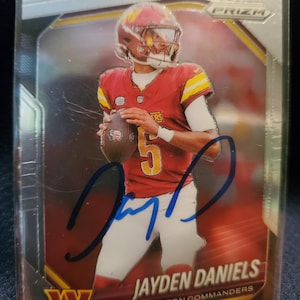 May include: A Panini Prizm trading card featuring Jayden Daniels of the Washington Commanders in a red and yellow uniform, holding a football. The card includes a blue ink autograph across the centre and the text Jayden Daniels Washington Commanders.