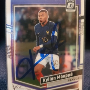 May include: A Donruss Optic trading card featuring a photograph of Kylian Mbappé in a dark blue France football shirt with the number 10, white shorts, and red socks. The card includes a blue ink autograph across the front and the FIFA logo.