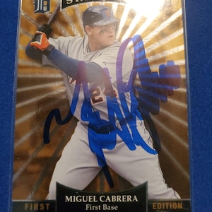 May include: A baseball trading card featuring Miguel Cabrera in a grey uniform with a Detroit Tigers logo, holding a bat. The card has a gold sunburst background, a blue ink autograph across the center, and text reading STARQUEST, FIRST EDITION, and First Base.