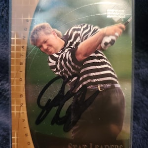 May include: A John Daly "Stat Leaders" trading card. The card features a photo of a golfer in a black and white striped shirt mid-swing. The card is signed and has the words "Driving Distance" on the left side.