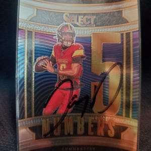 May include: A 2025 Select trading card featuring Jayden Daniels in a red Commanders uniform holding a football. The card displays the text Select, Numbers, Commanders, and Jayden Daniels, with a large gold number 5 and a black ink autograph.