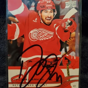 May include: A hockey trading card featuring Dylan Larkin of the Detroit Red Wings in a red uniform, celebrating with arms raised. The card has an autograph and the Upper Deck logo. The text "DYLAN LARKIN" and "DETROIT RED WINGS" are visible.