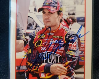 May include: A collectible trading card featuring a photograph of Jeff Gordon wearing a red, yellow, and blue racing suit and a matching cap. The card includes a blue ink autograph across the center and the text JEFF GORDON and TRAKS NASCAR NEXTEL CUP SERIES.