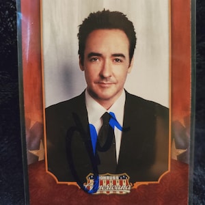 May include: A trading card featuring John Cusack in a suit and tie. The card has a red and gold border with the text "JOHN CUSACK" at the top. The card is signed with a blue pen and has the "Donruss Americana" logo.