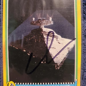 May include: A vintage trading card featuring a model Star Destroyer from Star Wars. The card has a yellow border with a blue and white banner at the bottom that reads "MODEL STAR DESTROYER". The card has a signature in purple ink.