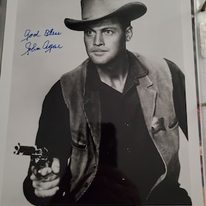 Peut inclure: Photographie en noir et blanc de John Agar, portant un chapeau de cowboy et un gilet, tenant un revolver. L'image comprend le texte "JOHN AGAR" en bas et une signature manuscrite avec les mots "God Bless" en haut.