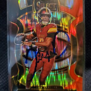 May include: A 2024 Select football trading card featuring Jayden Daniels in a burgundy and gold Washington Commanders uniform. The card has a holographic, reflective finish with an orange and gold background and a blue ink autograph across the front.