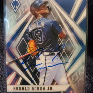 May include: A baseball trading card featuring Ronald Acuña Jr. in an Atlanta Braves uniform. The card has a blue and silver geometric design with an autograph in blue ink. The player is wearing a navy blue helmet and jersey with the number 13.