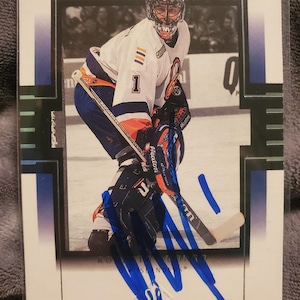 Puede incluir: Tarjeta de hockey que muestra a un portero con uniforme blanco, azul y naranja. El portero lleva un casco estampado y sostiene un palo de hockey. La tarjeta tiene un borde blanco y detalles azules.