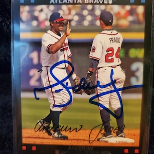 May include: A Topps Atlanta Braves baseball card featuring two players in their uniforms. One player is giving a high five. The card has the players' signatures in blue ink. The card is in a protective sleeve.