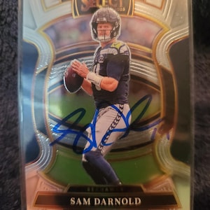 May include: A Sam Darnold football trading card. The card shows Darnold in a Seahawks uniform, holding a football. The card has a silver and gold border and is in a protective sleeve. The card is autographed in blue ink.