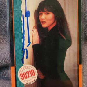 May include: A trading card featuring a woman with dark hair, wearing a black top, and leaning against a wall. The card has an orange border and a 90210 logo. Blue marker writing is visible on the card.