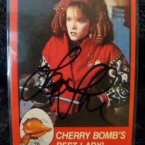 May include: A trading card featuring a woman in a red jacket with a signature. The card has a red border and includes the text "CHERRY BOMB'S BEST LADY!" and an illustration of a duck emerging from an egg.