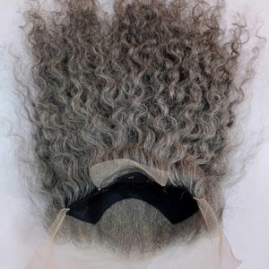 May include: A gray curly human hair wig with a lace front. The wig is laid flat on a white surface. The lace front is visible and the wig is ready to be styled.