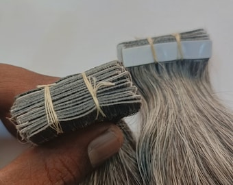 May include: A pack of gray human hair extensions with tape wefts. The wefts are stacked and secured with rubber bands.