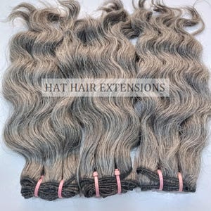 May include: Three bundles of wavy grey human hair extensions. The hair is tied together with black elastic bands and secured with pink plastic clips. The bundles are labelled "HAT HAIR EXTENSIONS".