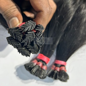 May include: Close-up of black hair extensions, with a bundle of the extensions held in a hand. The extensions are secured with red bands. The hair appears to be straight and sleek.