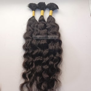 Può includere: Tre pacchetti di extension per capelli umani ondulati neri con elastici gialli in cima. I capelli sono legati insieme con un elastico giallo. Il testo "HAT HAIR EXTENSIONS" è visibile nell'immagine.